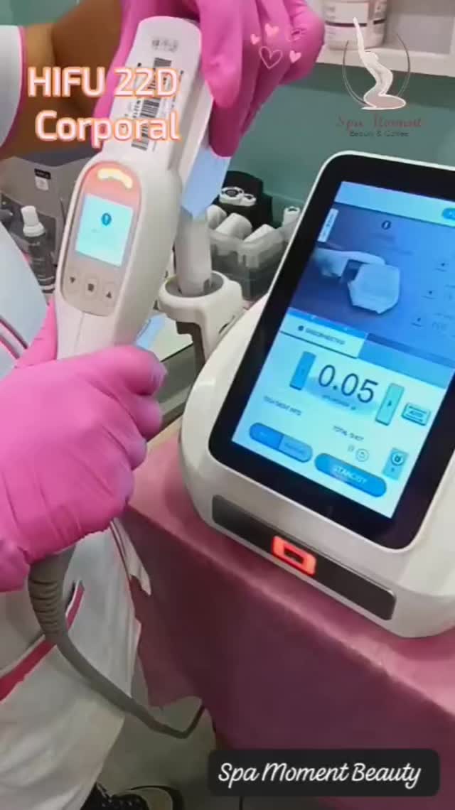 Manos con guantes rosas sostienen el cabezal del equipo de HIFU 22D frente a su pantalla, en la cabina de pared verde agua