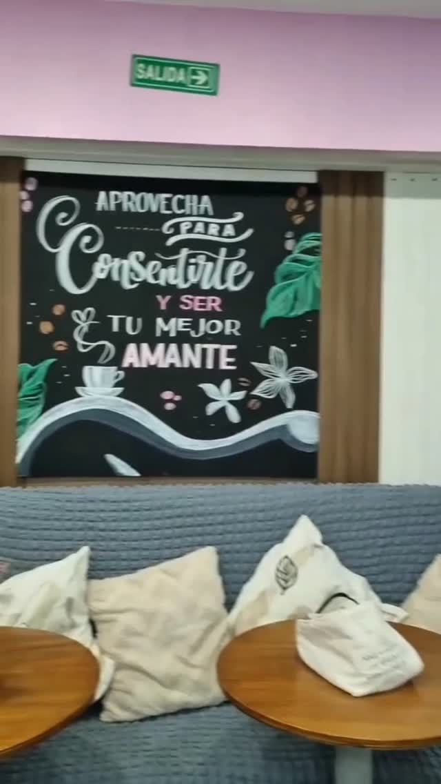 El rincón de café: una pizarra pintada a mano que dice «Aprovecha para consentirte», un sillón gris con almohadones y mesitas redondas de madera